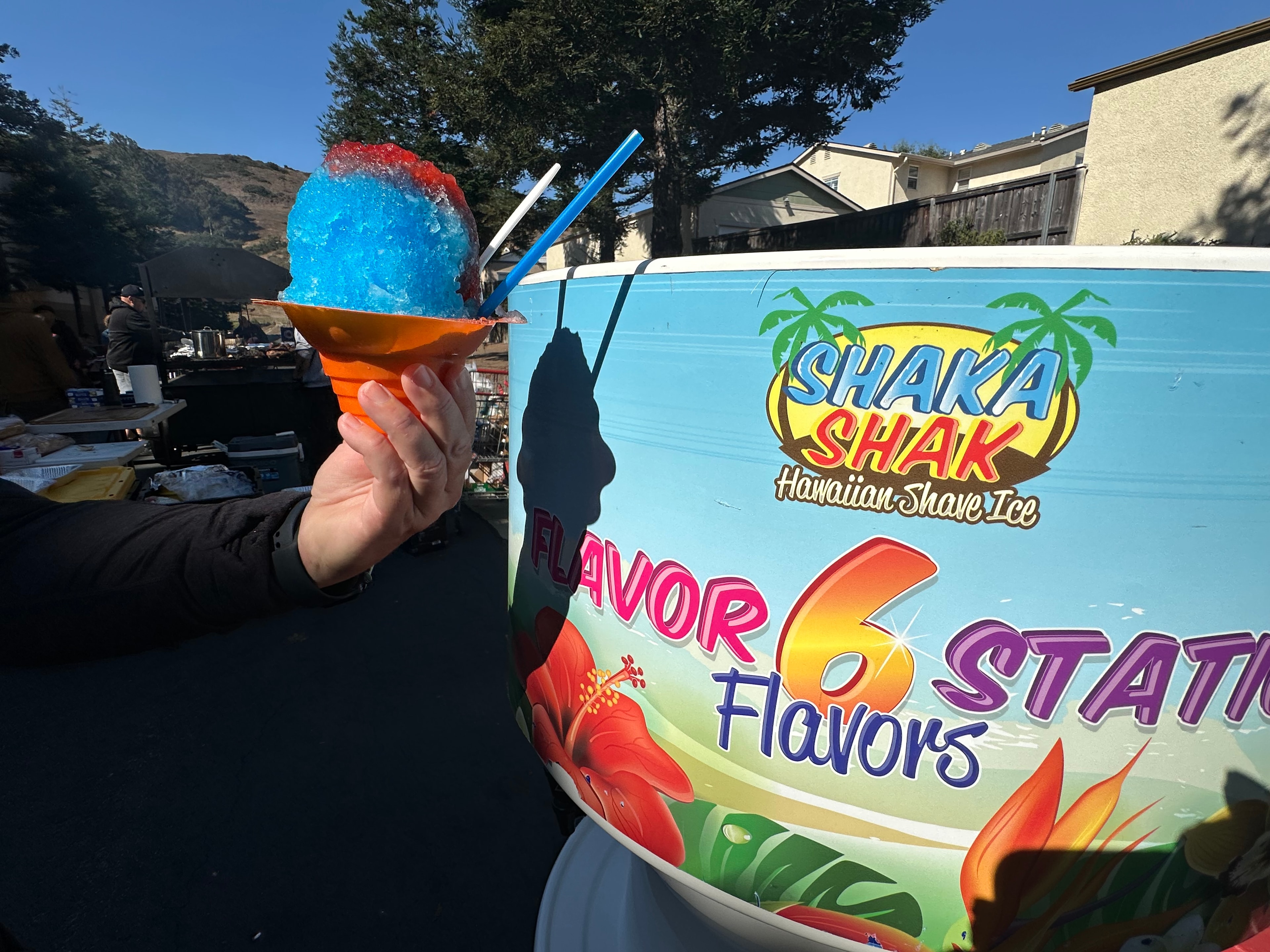 Hand holding a blue and red shave ice in front of the Shaka Shak flavor station