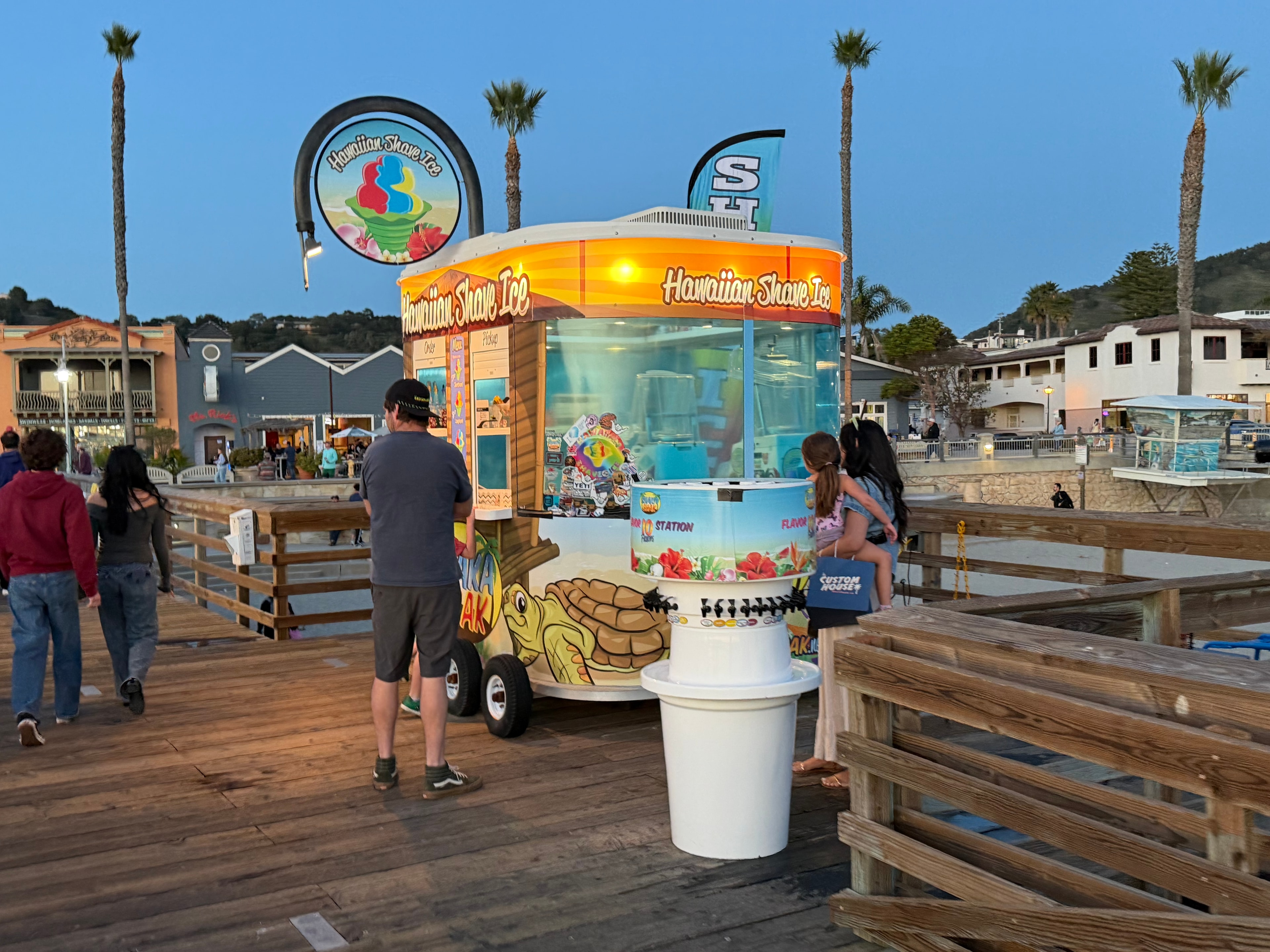 Shaka Shak kiosk on the pier at dusk with palm trees and town in background