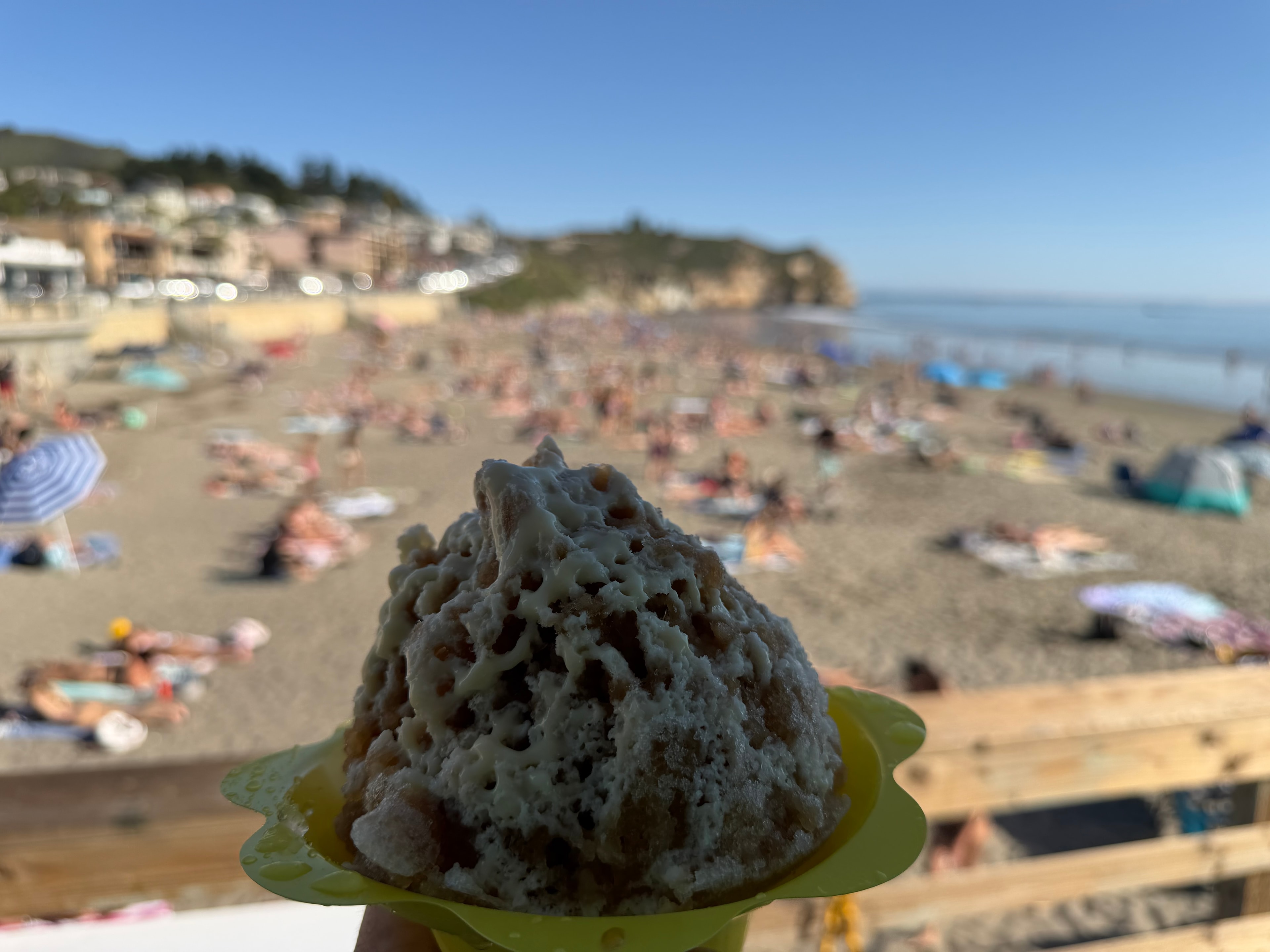 Shave ice in a Shaka Shak cup held up with a packed Avila Beach in the background