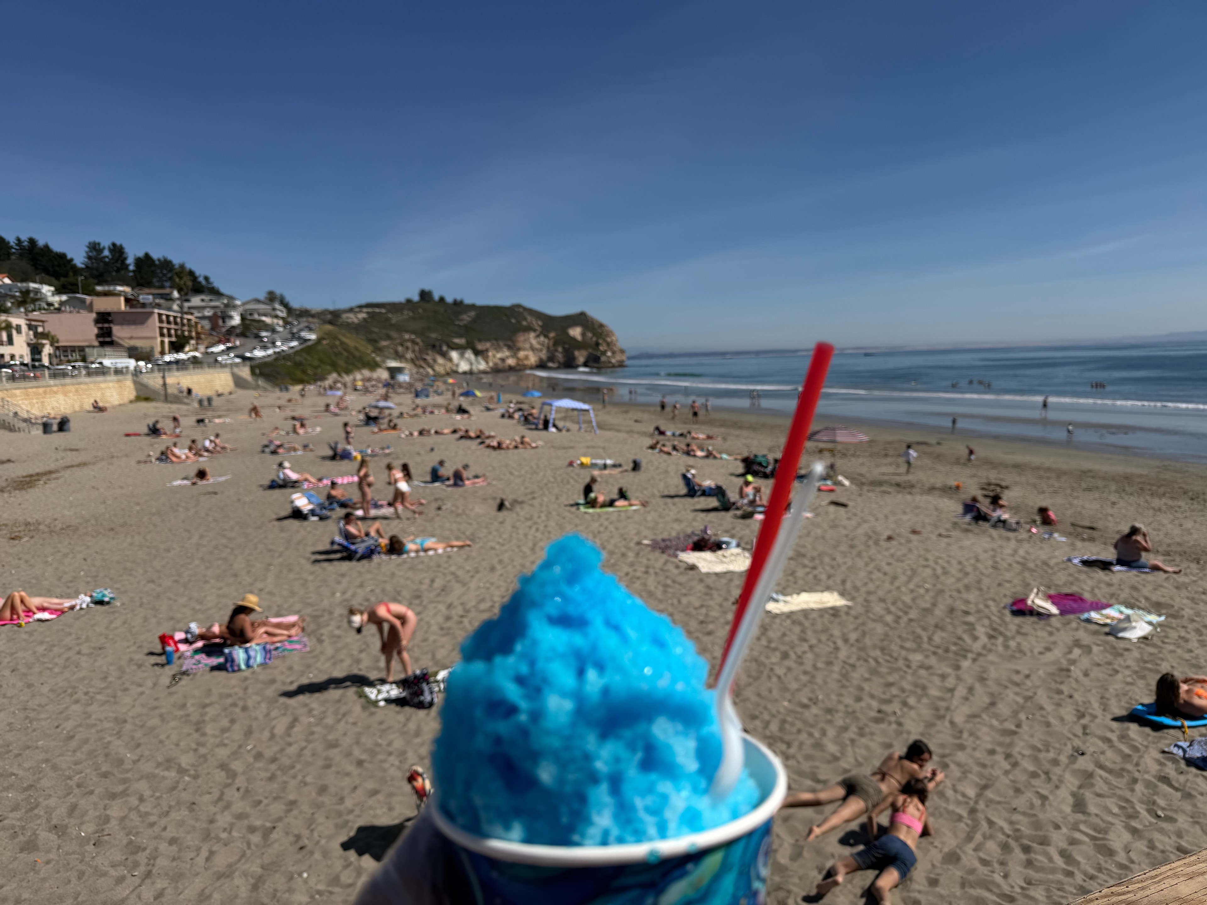 Blue shave ice in a Shaka Shak cup held up overlooking a packed Avila Beach