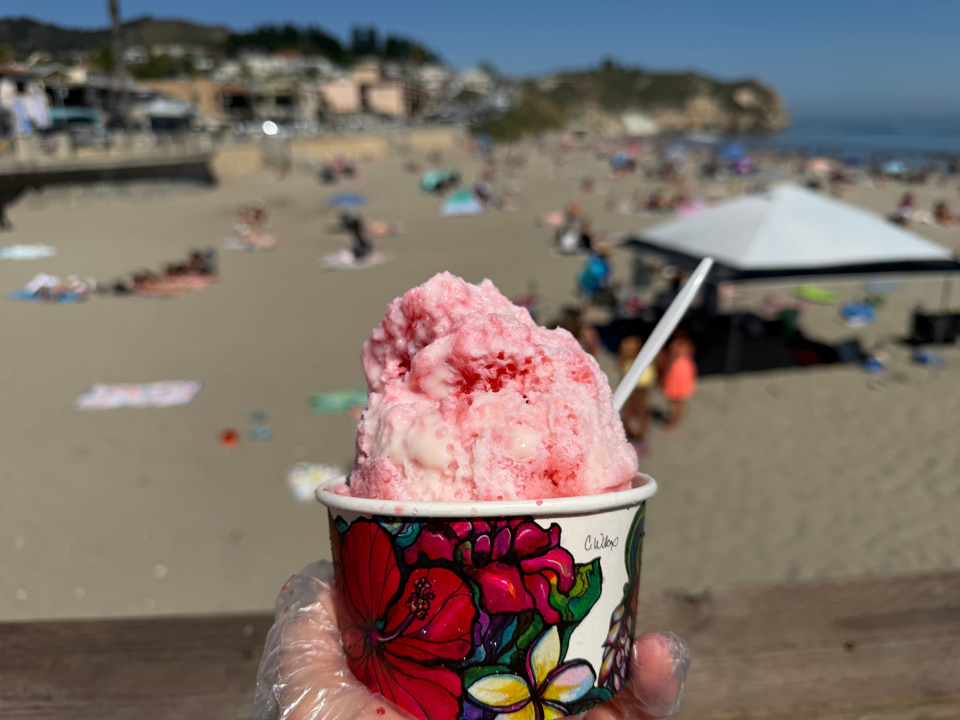 Pink strawberry shave ice in a floral cup held up over a packed Avila Beach
