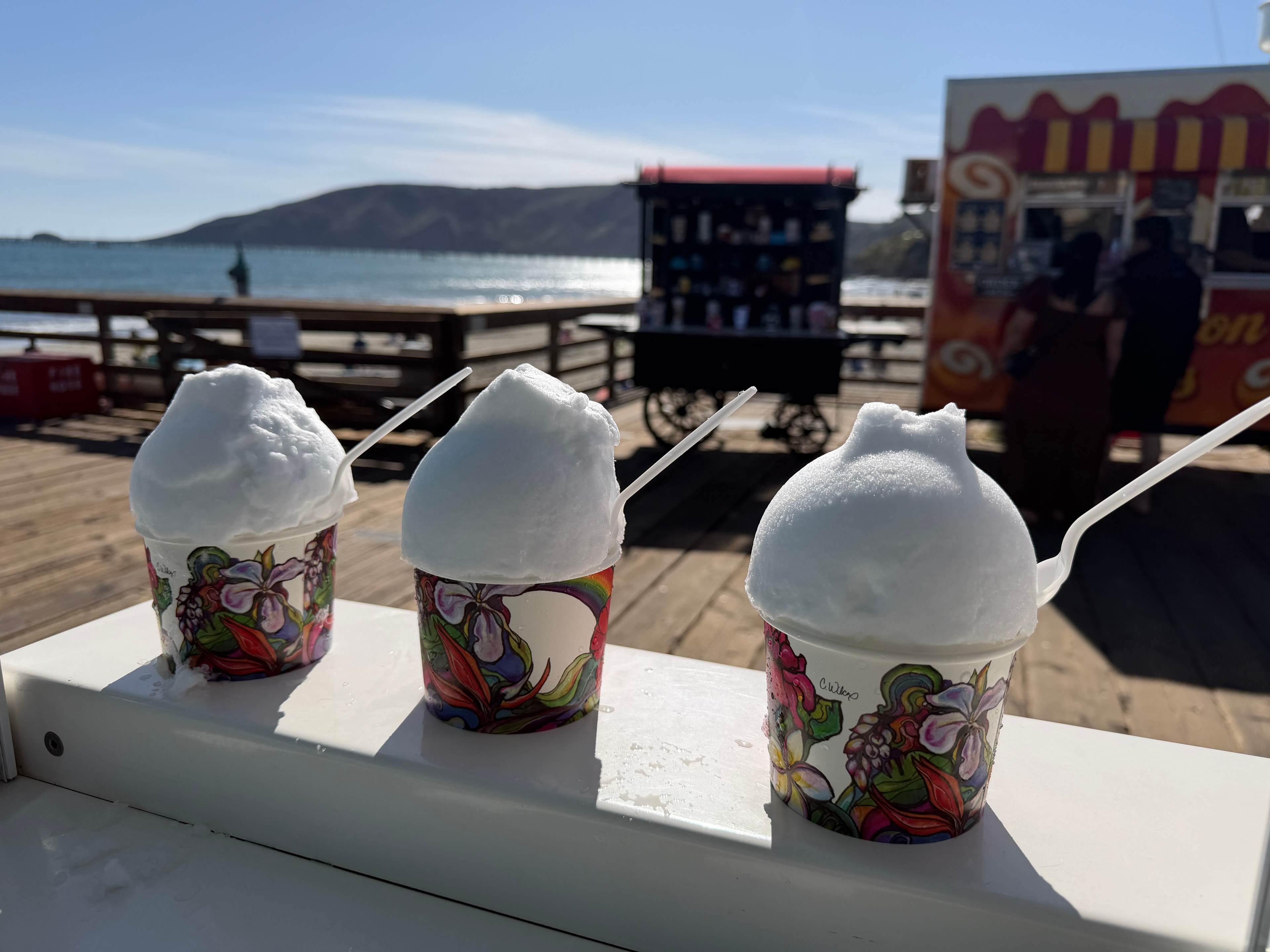 Three shave ice cups in floral Shaka Shak cups lined up on the pier railing