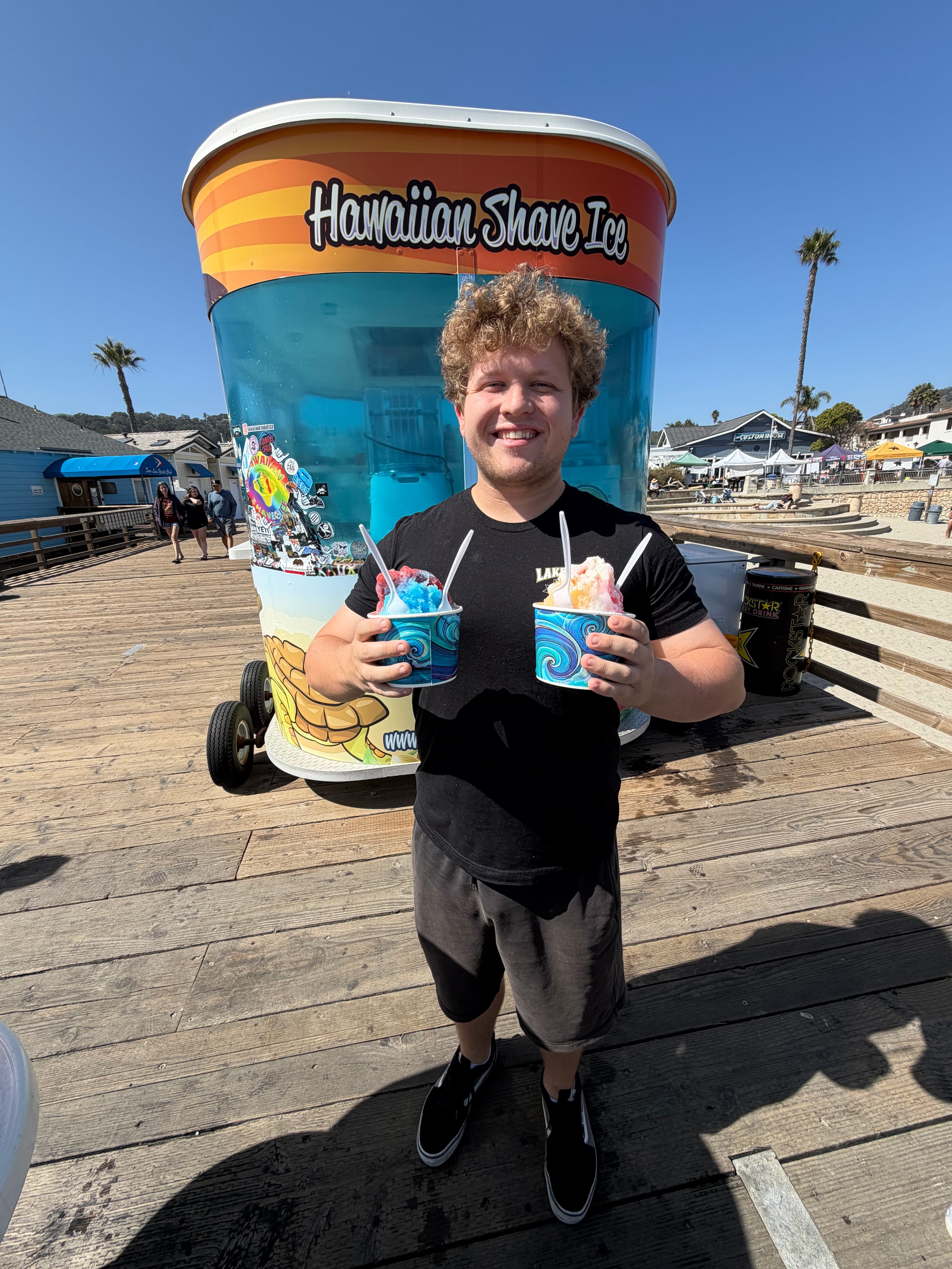 Guy holding two shave ice cups posing through the giant Hawaiian Shave Ice cup cutout on the Avila Beach pier